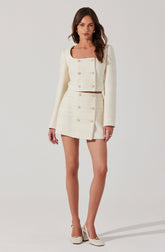 A woman is wearing a cream-colored, textured, cropped blazer with long sleeves and a square neckline, paired with a matching high-waisted mini skirt featuring double-breasted gold buttons down the front. She is also wearing cream-colored heels with a strap and a decorative bow.